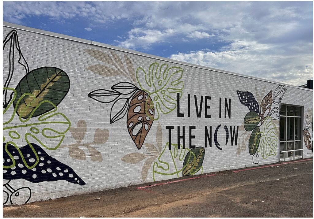 A mural on a white brick wall with botanical designs that says Live In The Now