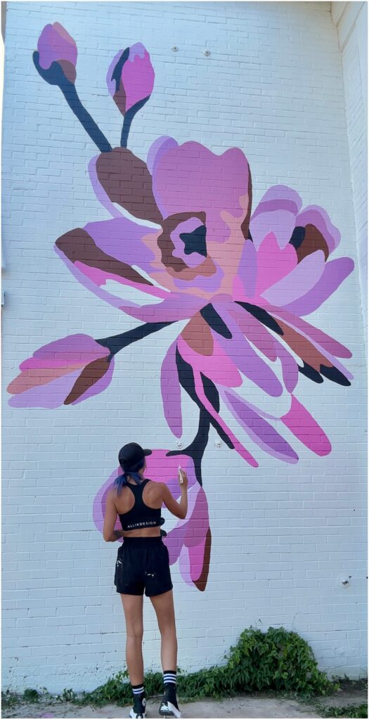 Texas muralist Alli K of Alli K Design painting a pink floral mural on a white brick wall in Dallas